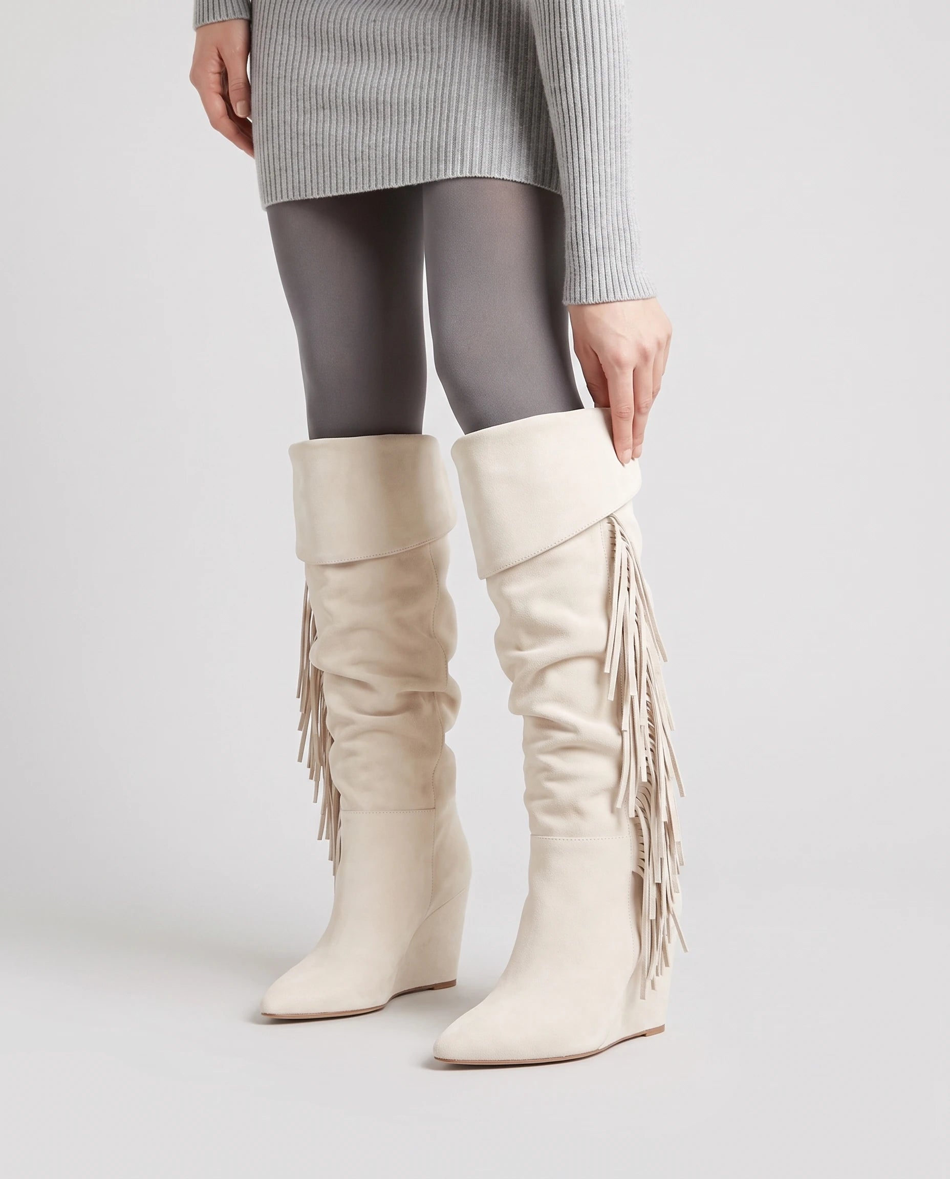 Bottes hautes à franges à talon compensé Alexandrine — Blanc Cassé, portées en plan rapproché, main sur le revers