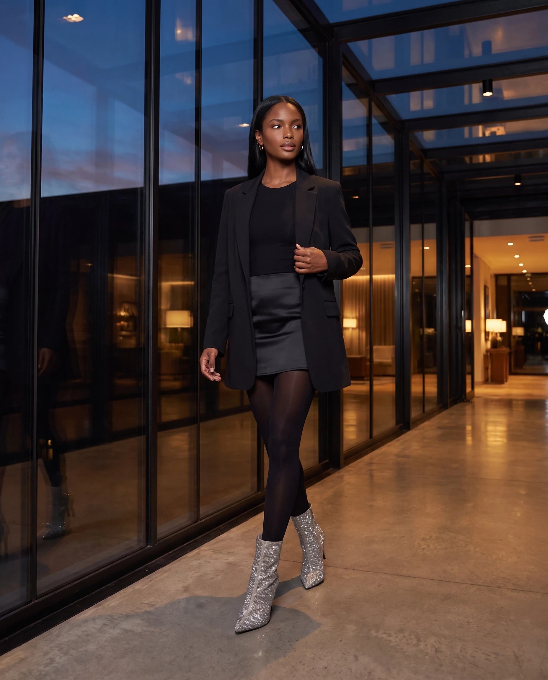 Bottines à strass à talon aiguille Perrette — Argent, portées dans un passage vitré d'hôtel au crépuscule