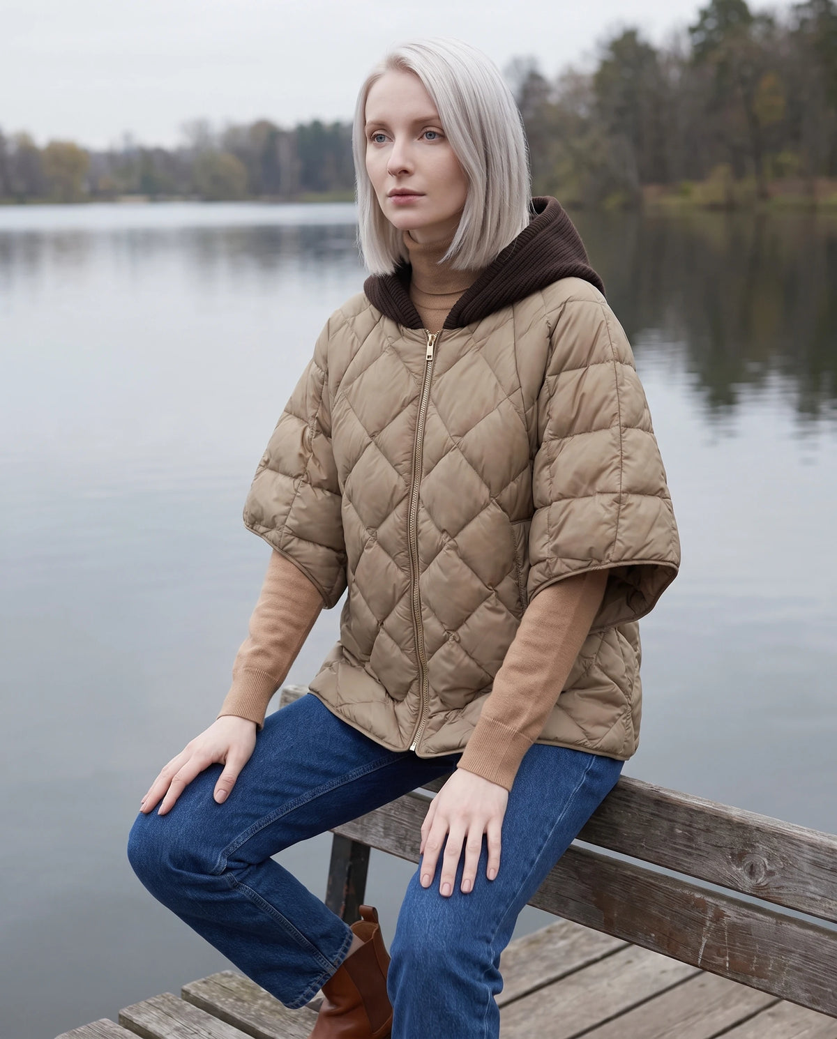 Cape matelassée à capuche tricot Stella en Sable, portée sur un banc de bois face à un lac
