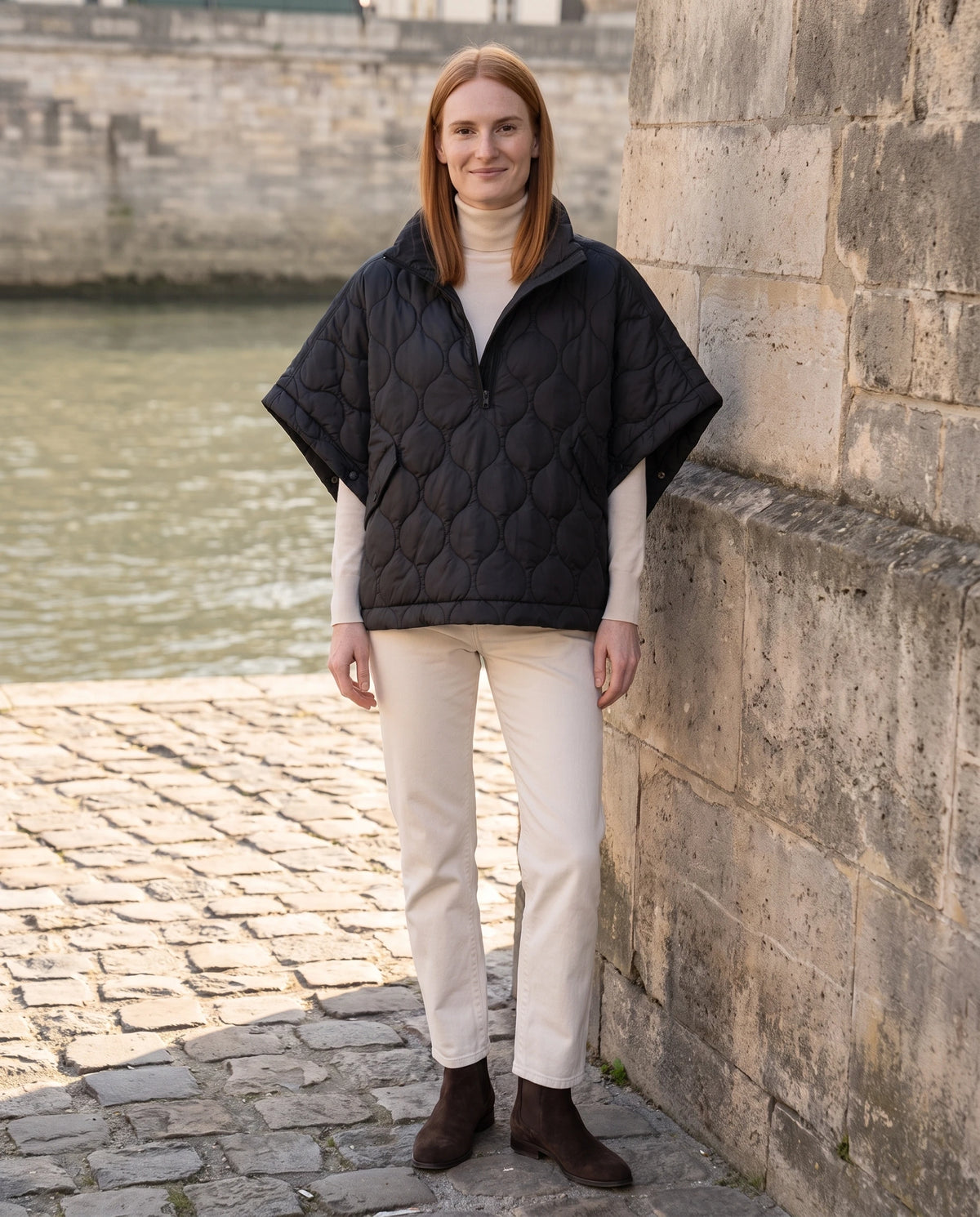 Cape matelassée à demi-zip Orane en Noir, portée sur un quai pavé au bord de l'eau, demi-zip ouvert