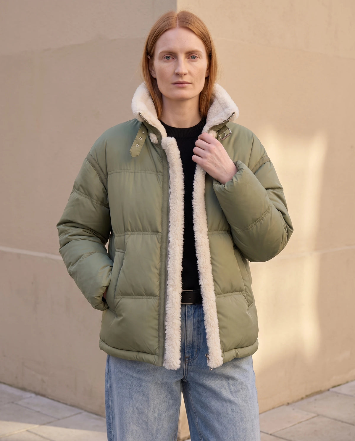 Doudoune courte oversize bordée sherpa Laurianne vert de gris, mur beige en fin de journée, main au col