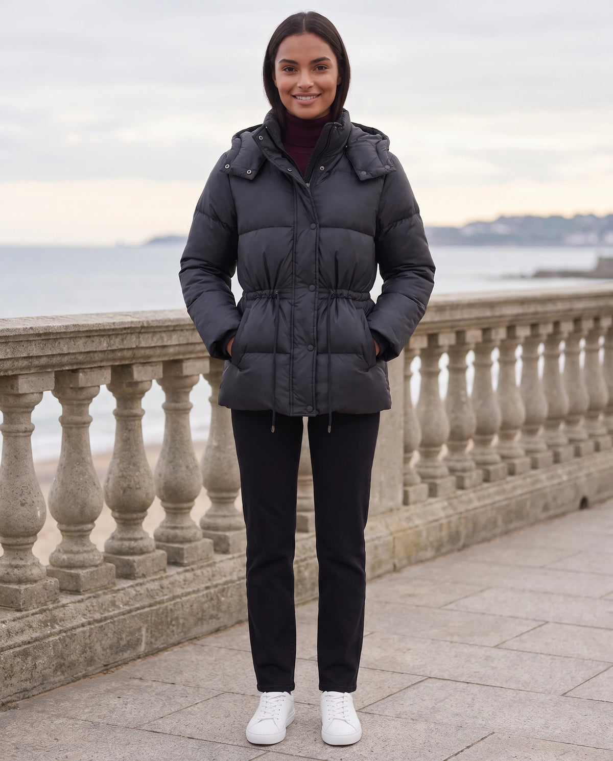 Kurze Steppjacke mit Kapuze und elastischer Taille Annick in Schwarz, getragen auf einer Promenade am Meer