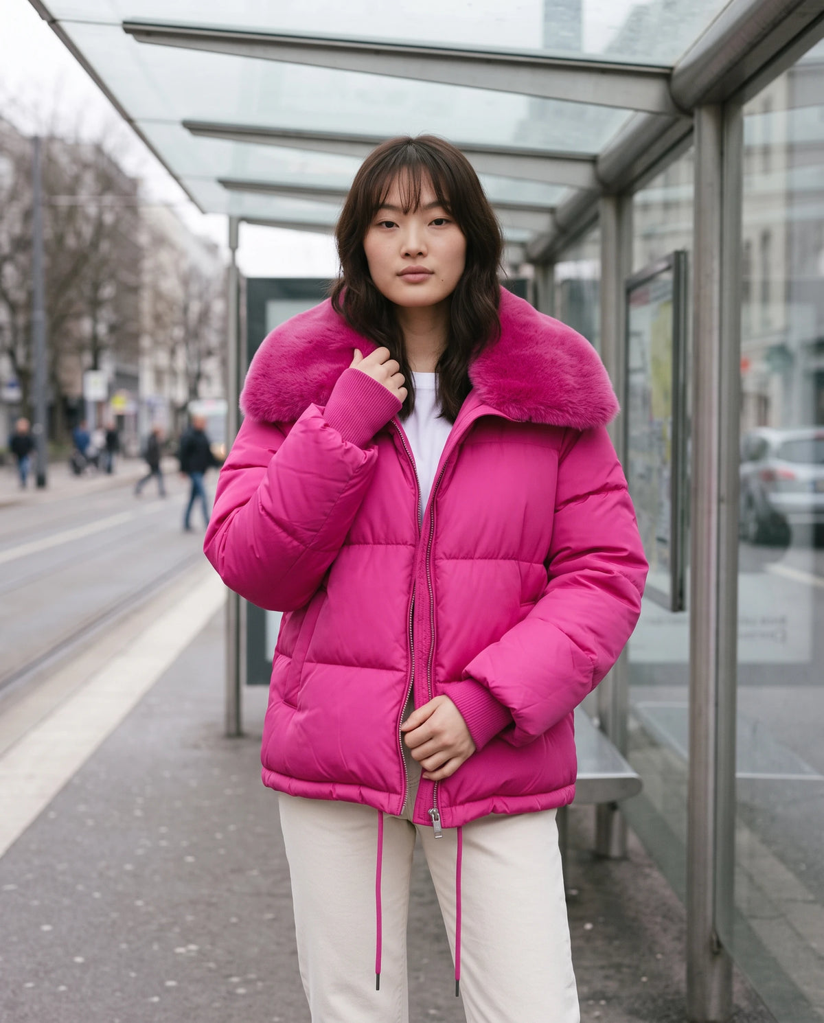 Kurze Steppjacke mit grossem Kunstfellkragen Mila in Fuchsia, getragen unter dem Glasdach einer Tramhaltestelle ohne Plakat