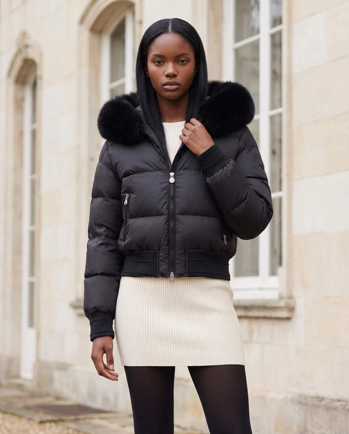 Kurze Steppjacke mit grosser Kunstfellkapuze Rebecca in Schwarz · schwarzes Fell, getragen vor der Steinfassade eines Stadtpalais