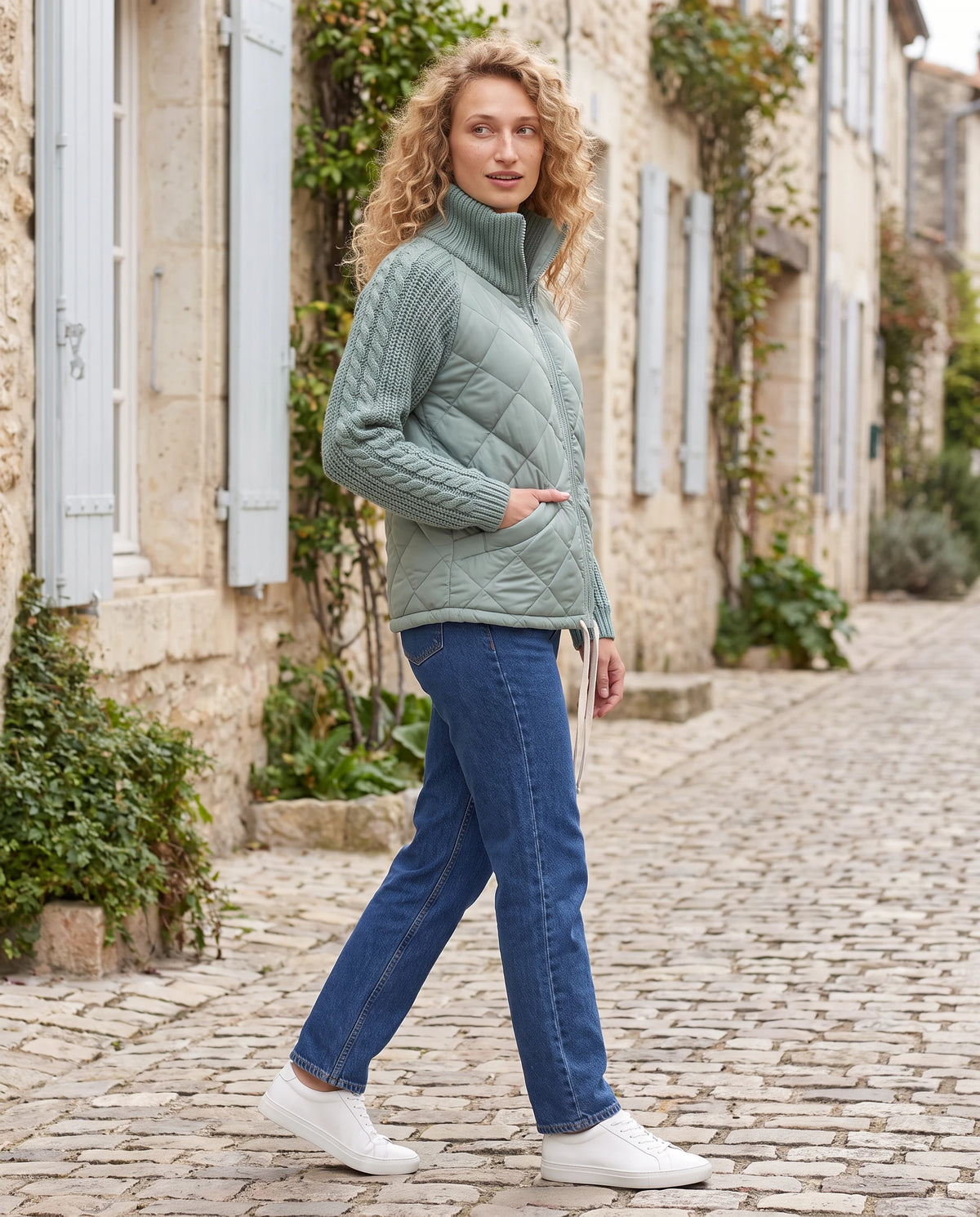 Doudoune courte à manches tricot Emma en Vert Sauge, portée dans une ruelle pavée aux volets pâles