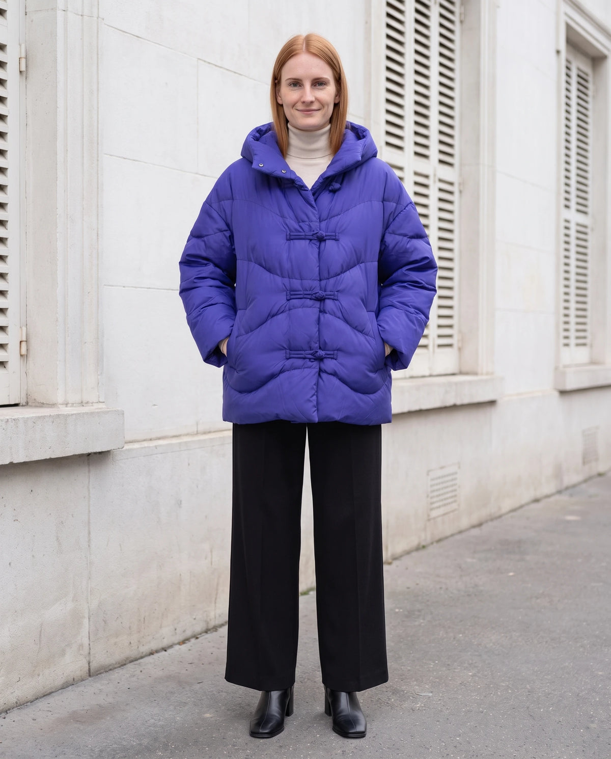 Doudoune courte oversize à capuche et brandebourgs Andréa en Bleu Roi, portée devant une façade de pierre blanche