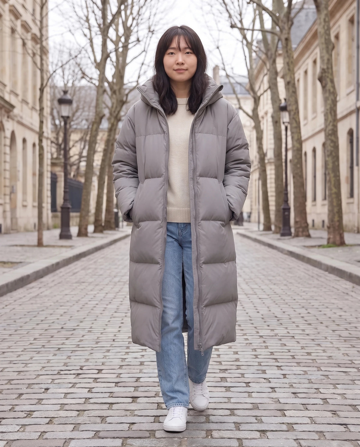 Doudoune extra-longue à capuche amovible Nolwenn en Gris, portée dans une rue pavée bordée de platanes nus, capuche retirée, col montant seul