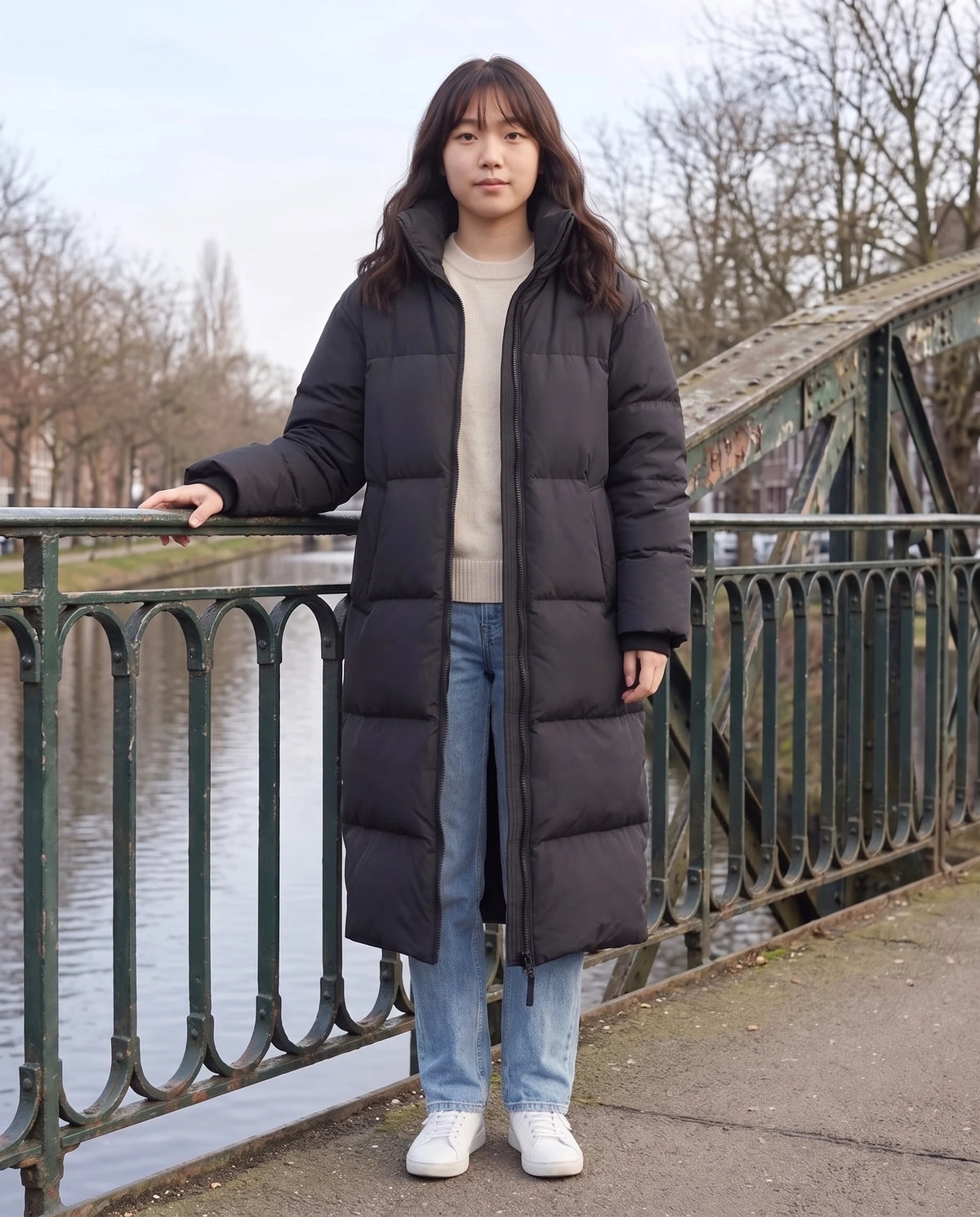 Doudoune extra-longue à capuche amovible Nolwenn en Noir, portée sur un pont de fer au-dessus d'un canal