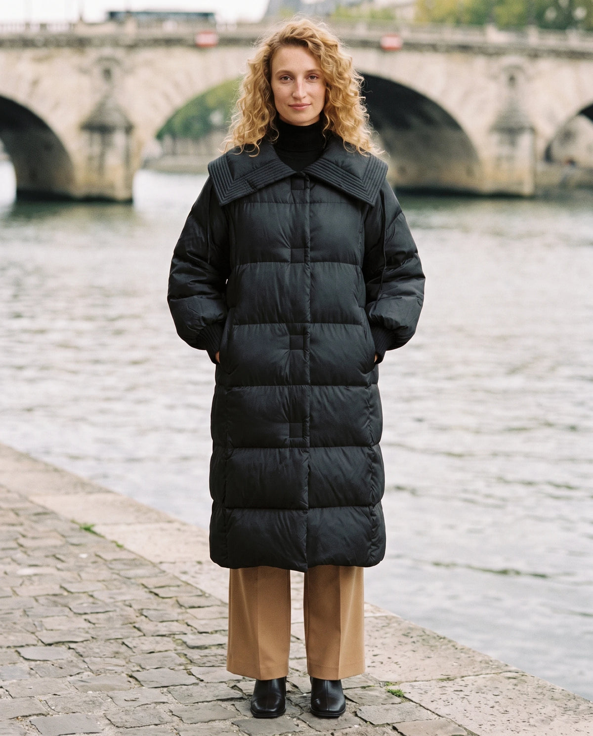 Doudoune extra-longue à grand col carré et poignets tricot Anastasia en Noir, portée sur un quai de pierre le long d'un fleuve