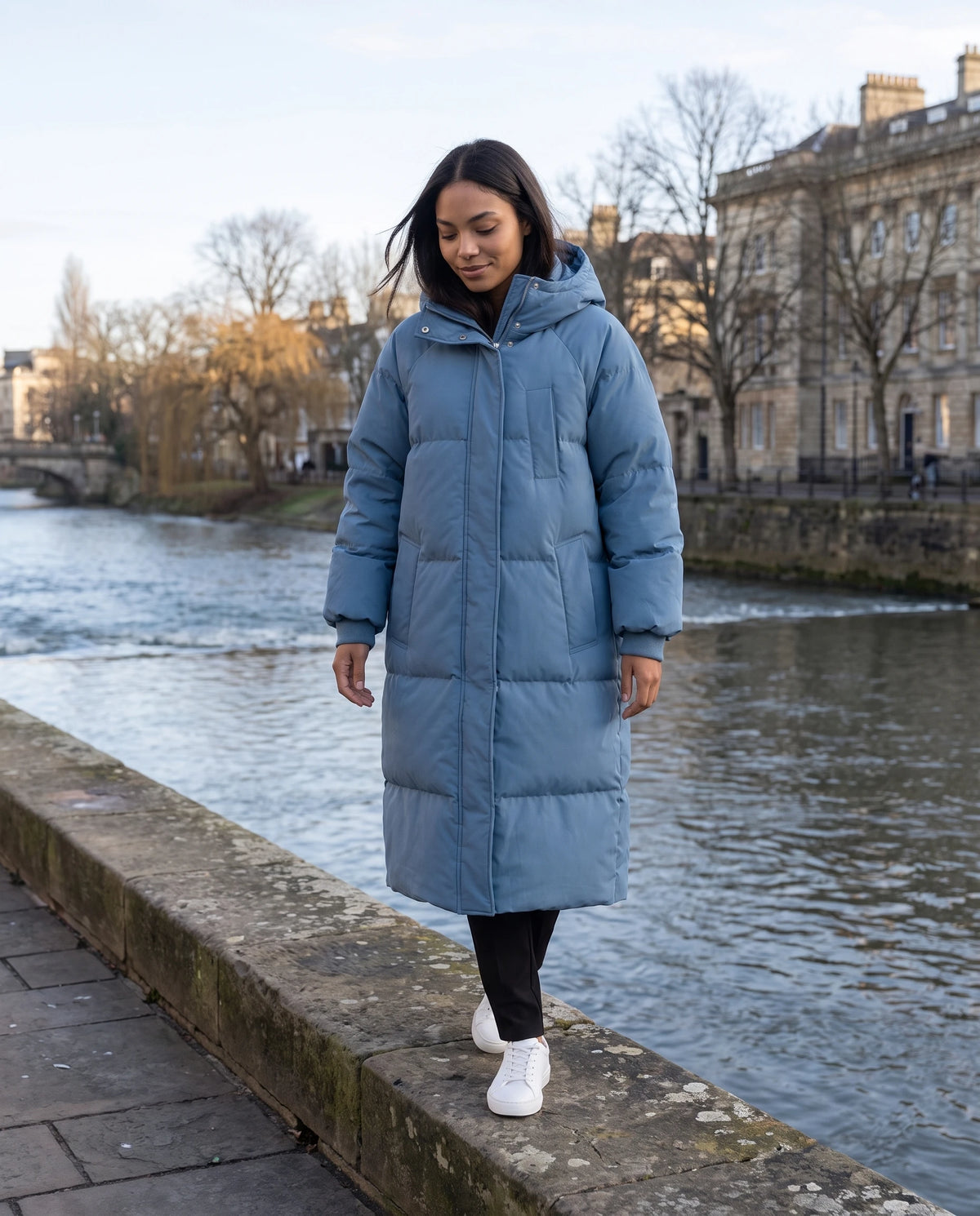 Doudoune longue à capuche Claire en Bleu Denim, portée le long d'un quai fluvial