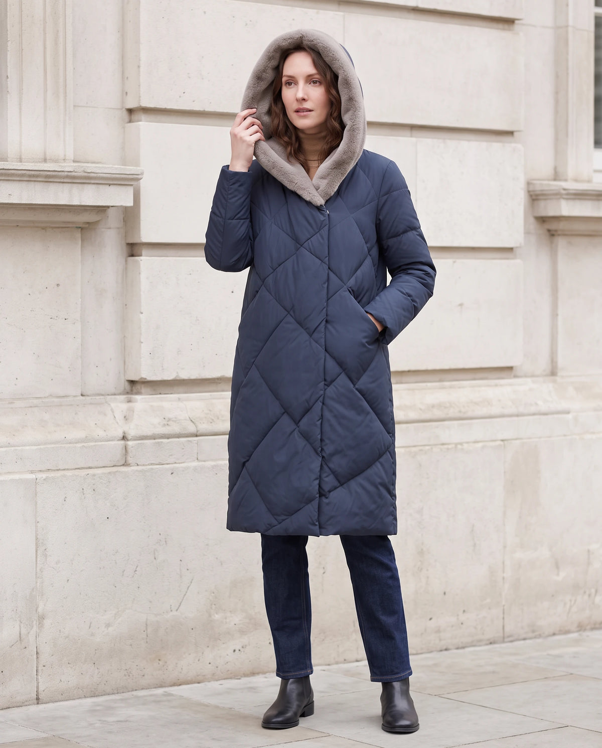 Lange Steppjacke mit kunstfellgefütterter Kapuze Nadia in Marineblau, getragen vor einer hellen Steinfassade, Kapuze aufgesetzt
