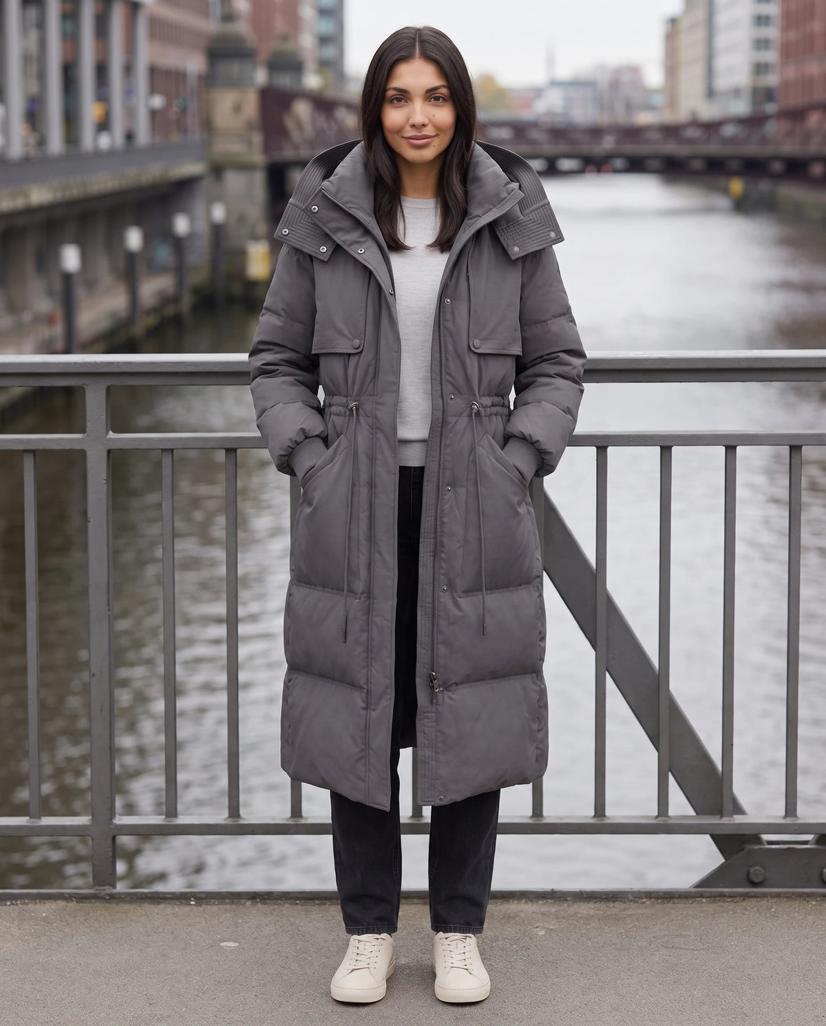 Lange Steppjacke mit Kapuze und Tunnelzugtaille Mélissa in Anthrazit, getragen auf einer Stahlbrücke in der Stadt