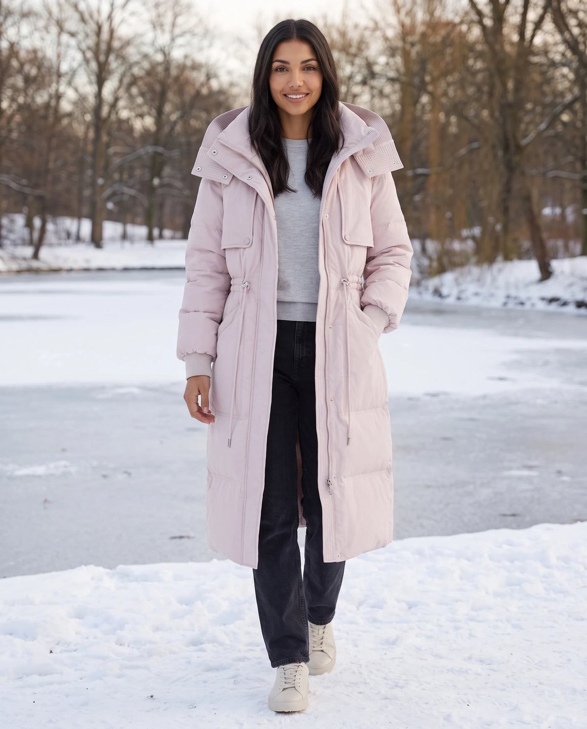 Lange Steppjacke mit Kapuze und Tunnelzugtaille Mélissa in Puderrosa, getragen in einem verschneiten Park an einem zugefrorenen Teich