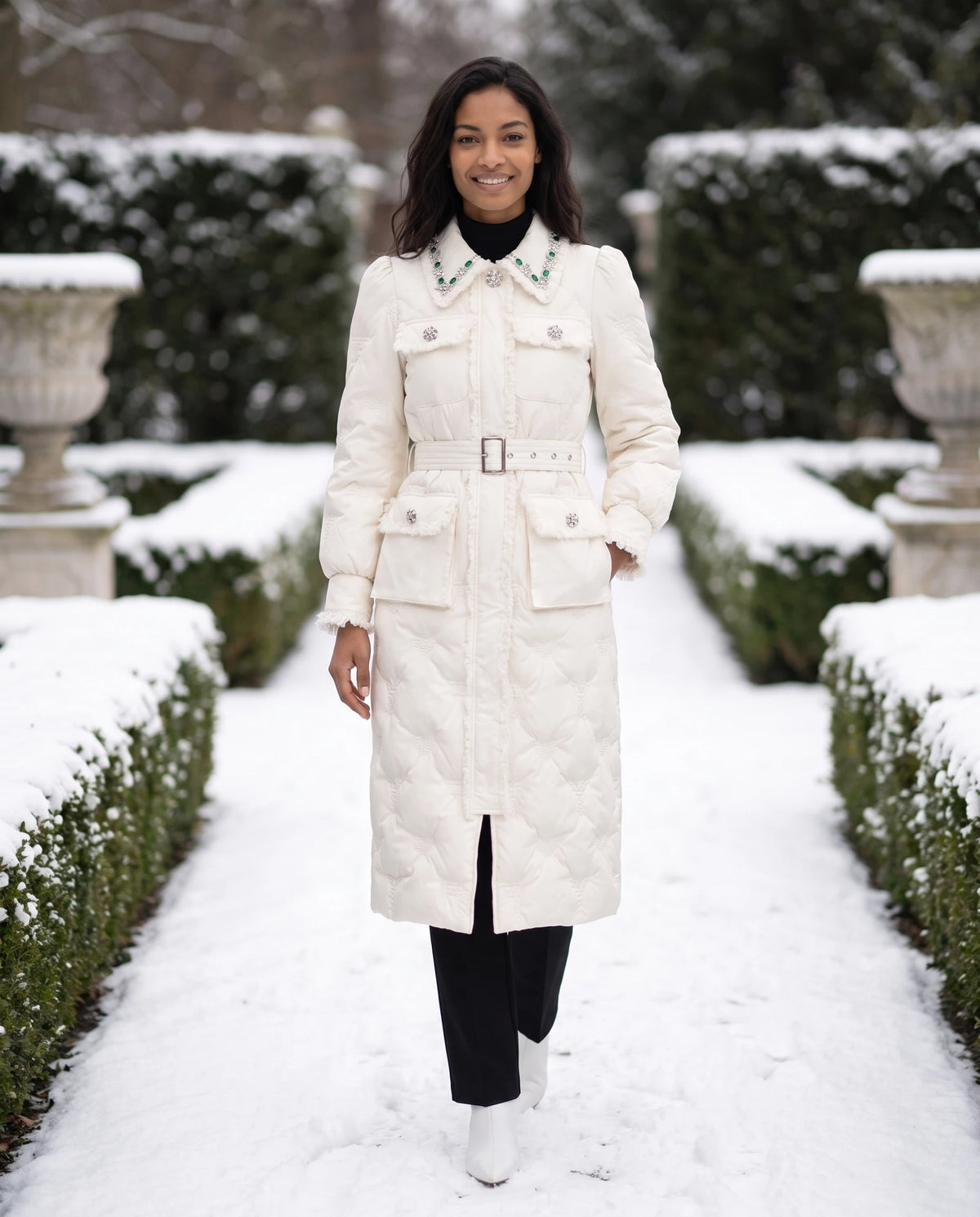 Doudoune longue ceinturée à col bijoux et manches bouffantes Déborah en Blanc Perle, portée dans une allée de jardin à la française enneigée