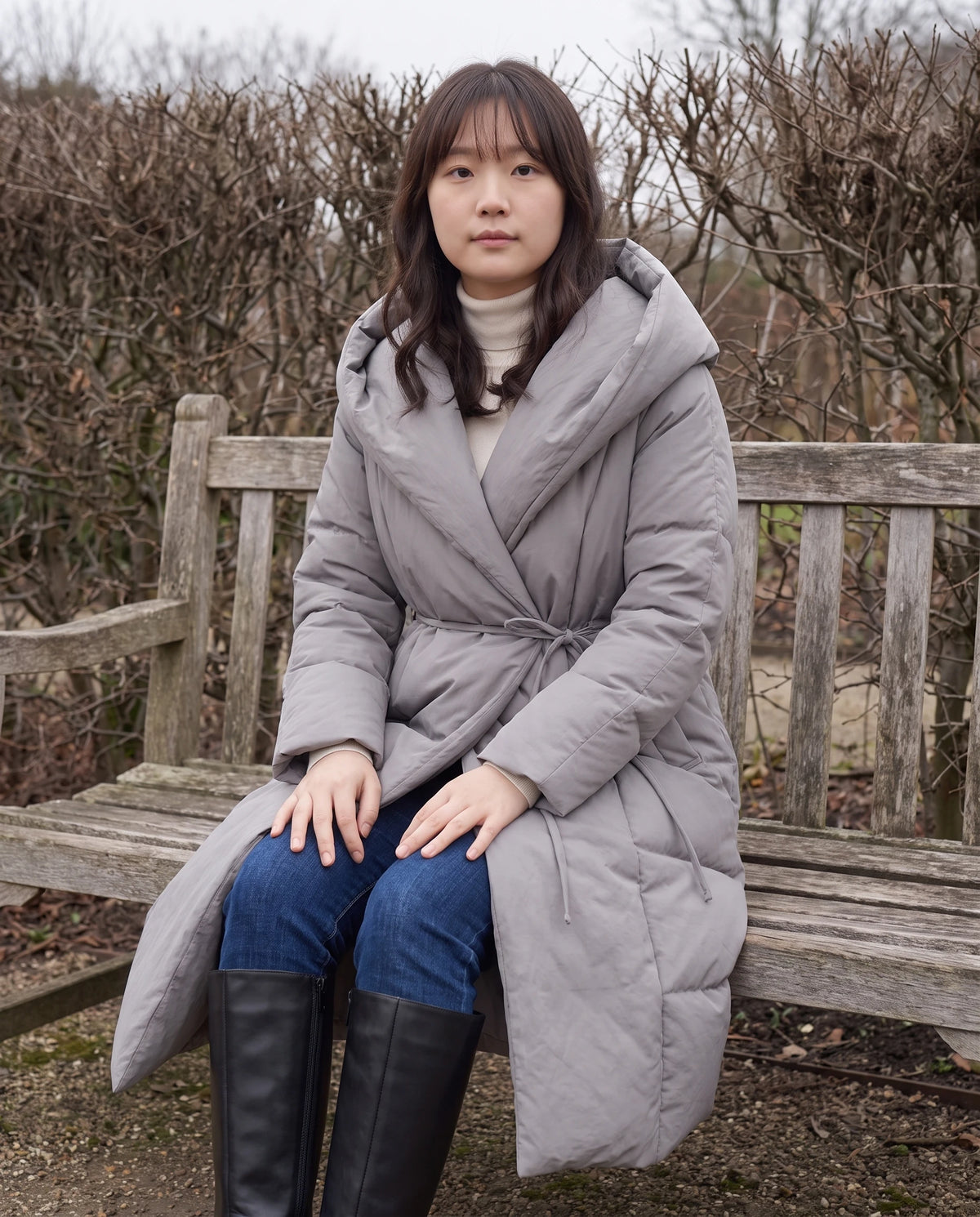Doudoune longue portefeuille à capuche oversize Aurélia en Gris, portée assise sur un banc de jardin en hiver