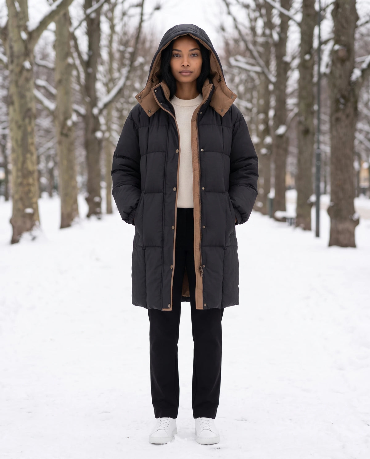 Mittellange Winterjacke mit Kapuze und Kontrastfutter Alexia in Schwarz, getragen mit Kapuze auf einer verschneiten Platanenallee