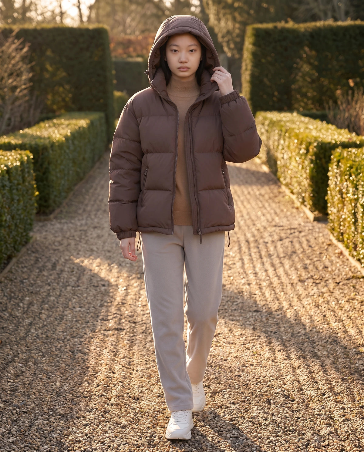 Doudoune courte oversize à capuche amovible Camélia, coloris Brun Cacao, capuche relevée dans une allée bordée de haies