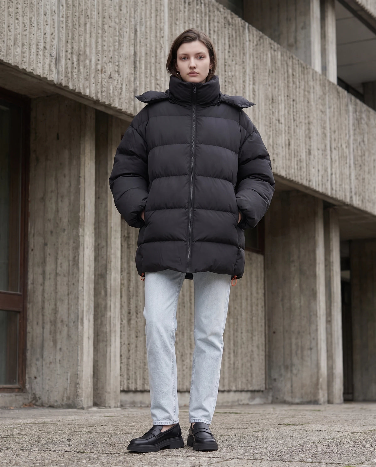 Oversize-Pufferjacke mit Kapuze Freya in Schwarz, getragen vor einem brutalistischen Gebäude