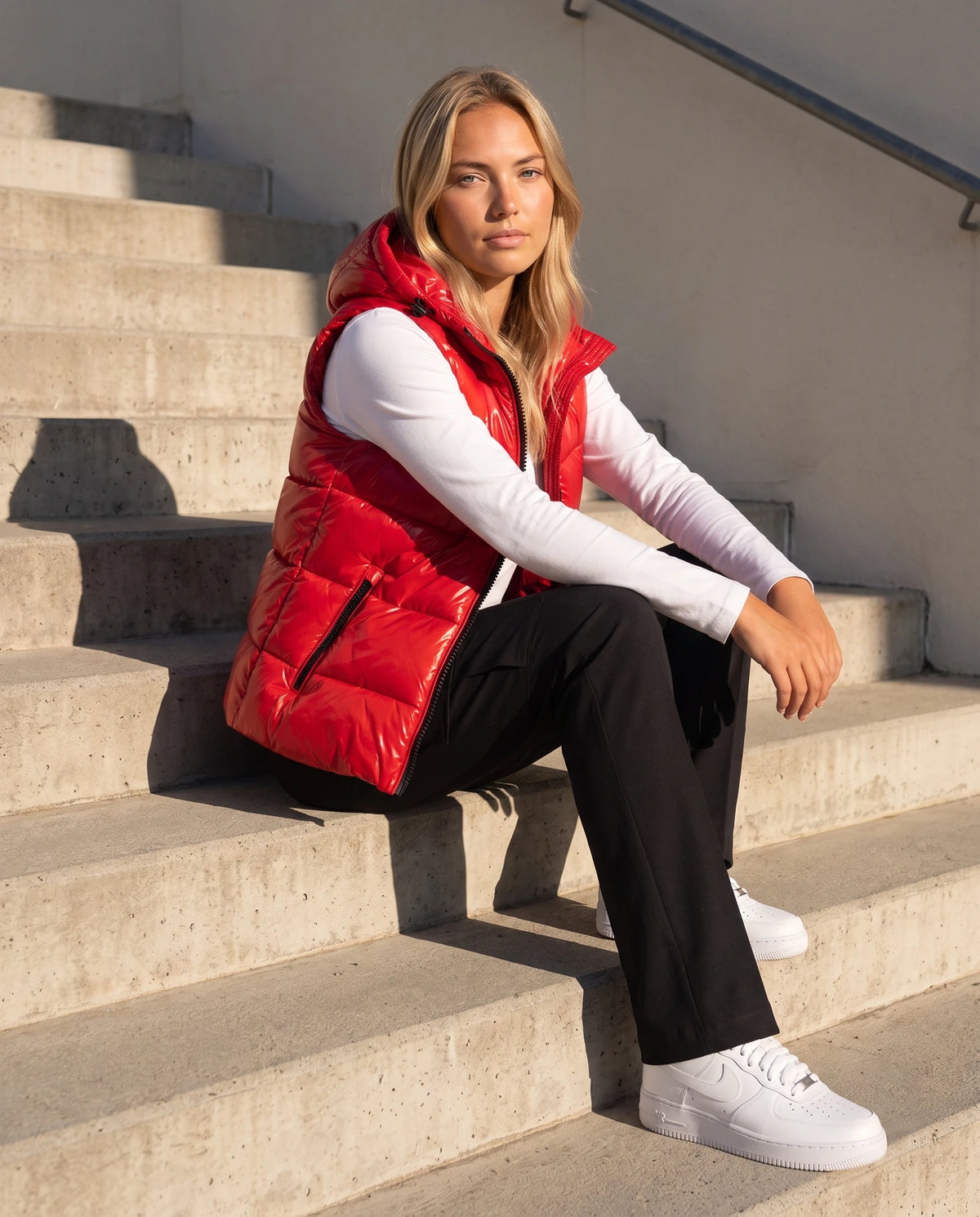 Doudoune sans manches à capuche métallisée Rosanne, coloris Rouge, assise sur un escalier en béton au soleil