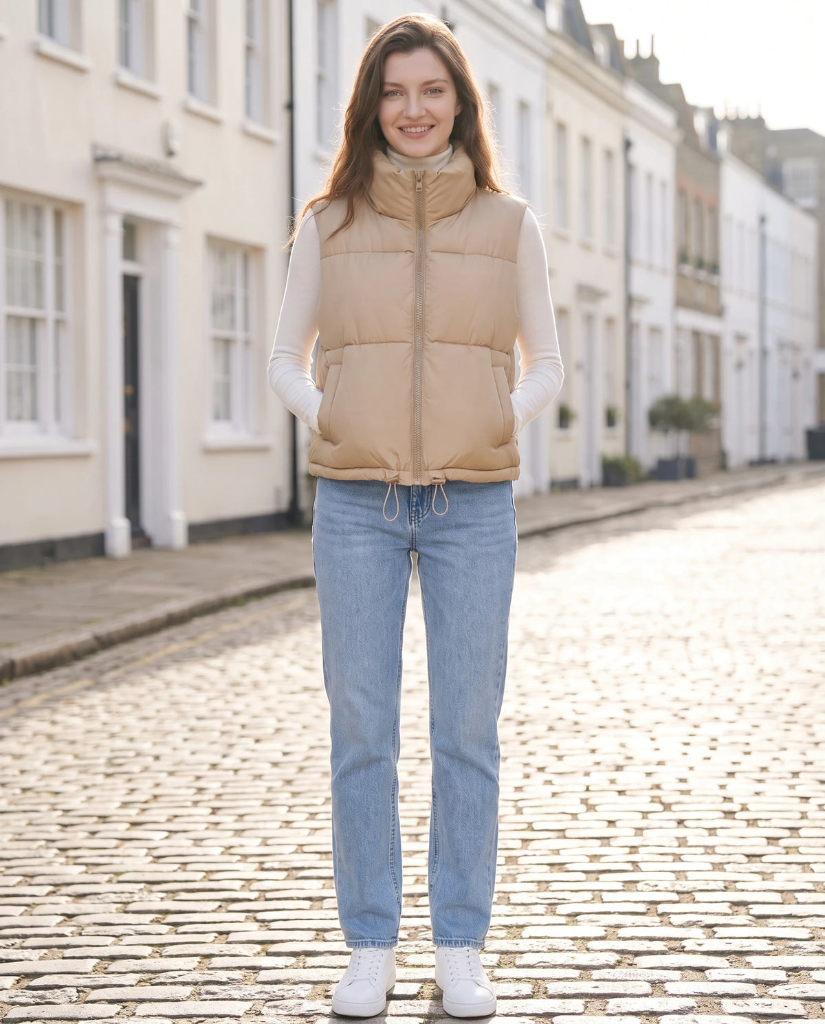 Gilet matelassé cropped à col montant et ourlet à cordon Lydie en Beige, portée dans une rue pavée bordée de maisons claires au soleil