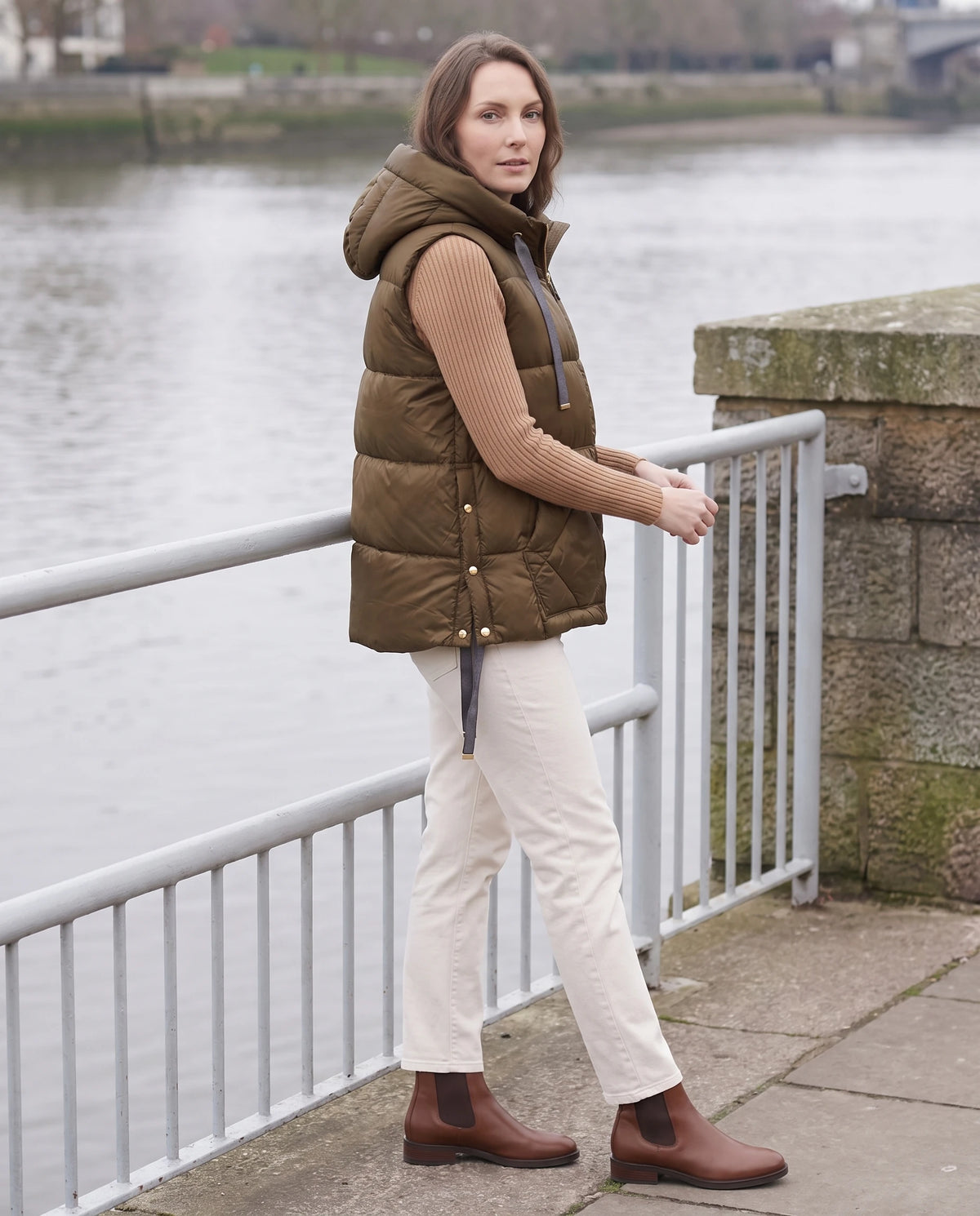 Gilet matelassé à capuche et grandes poches Corinne en Brun, porté sur un quai au bord de l'eau