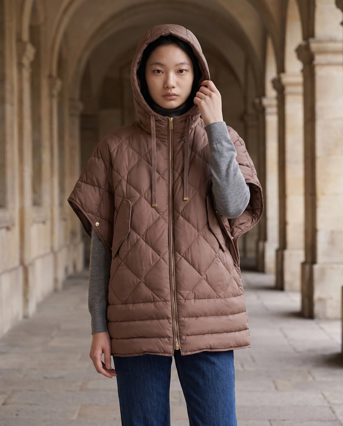 Gilet matelassé à capuche et pressions Céline en Brun Cacao, portée sous les arcades de pierre d'un bâtiment ancien, capuche relevée