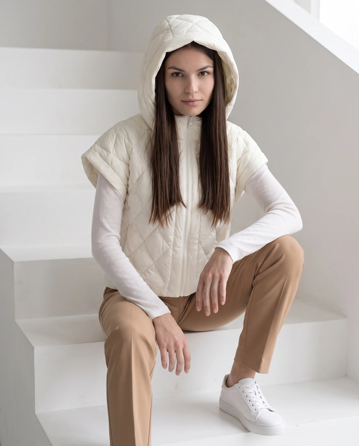 Gilet matelassé court à capuche Mia en Écru, portée sur l'escalier blanc d'un bâtiment moderne, capuche relevée