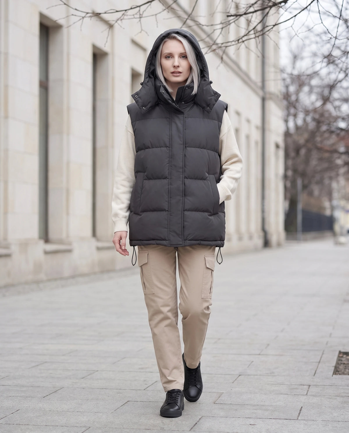 Gilet matelassé oversize à capuche amovible Patricia en Gris Anthracite, capuche relevée, sur un trottoir devant une façade en pierre claire