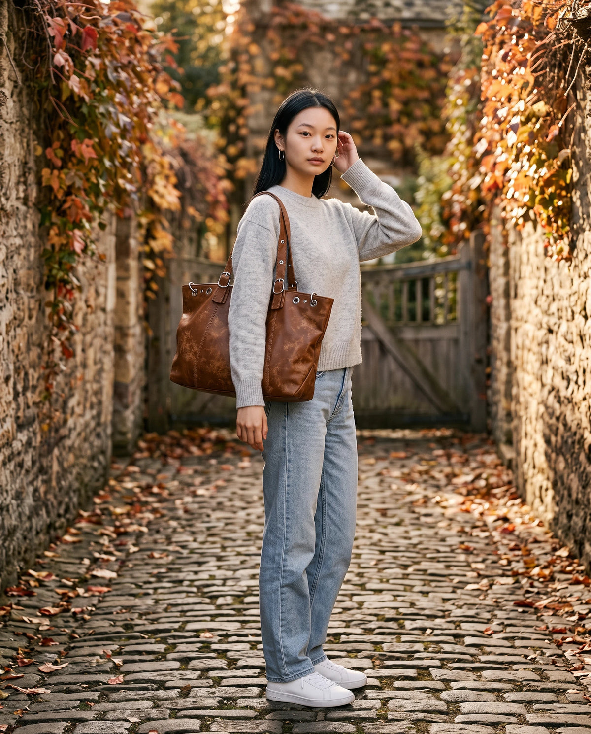 Große Hobo-Handtasche im Vintage-Look mit Ösen Billie in Cognacbraun, getragen in einer gepflasterten, efeubewachsenen Gasse