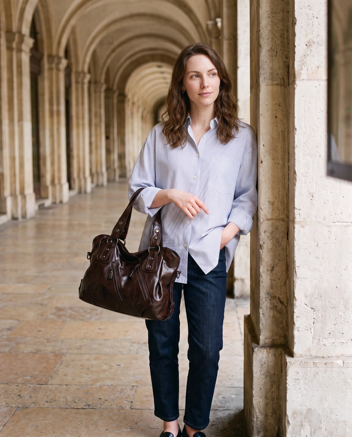Grand sac à main hobo bandoulière style motard à zips Thelma coloris Chocolat Foncé, porté au creux du bras sous une colonnade de pierre