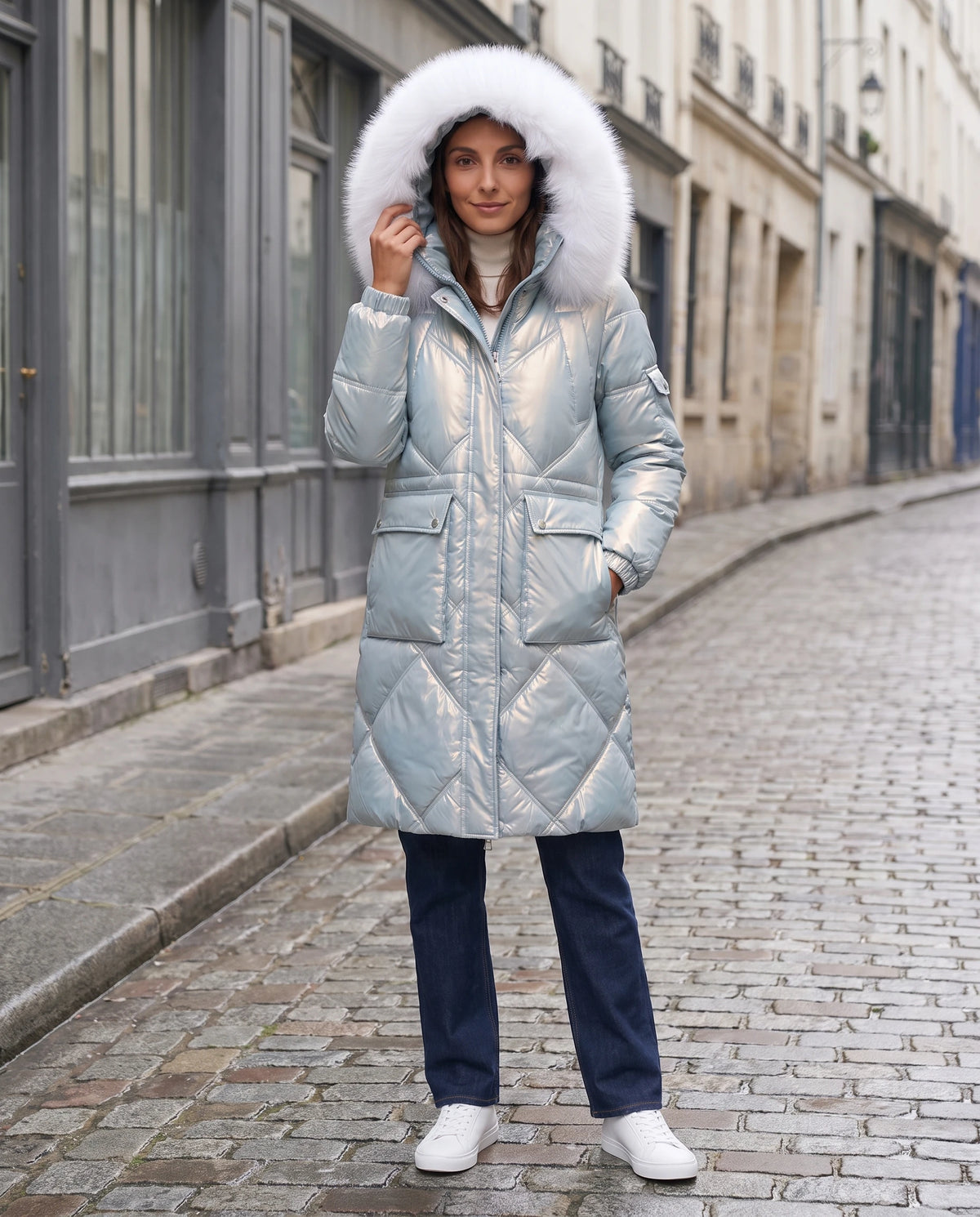 Parka mi-longue brillante à capuche fausse fourrure Vanessa en Bleu Ciel, portée dans une rue pavée aux façades grises, capuche relevée