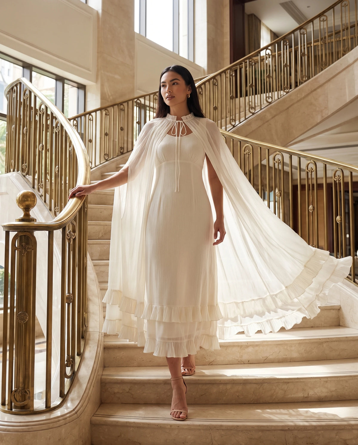 Robe longue cape en mousseline plissée Azalée en Ivoire, portée dans un escalier de marbre à rampe dorée