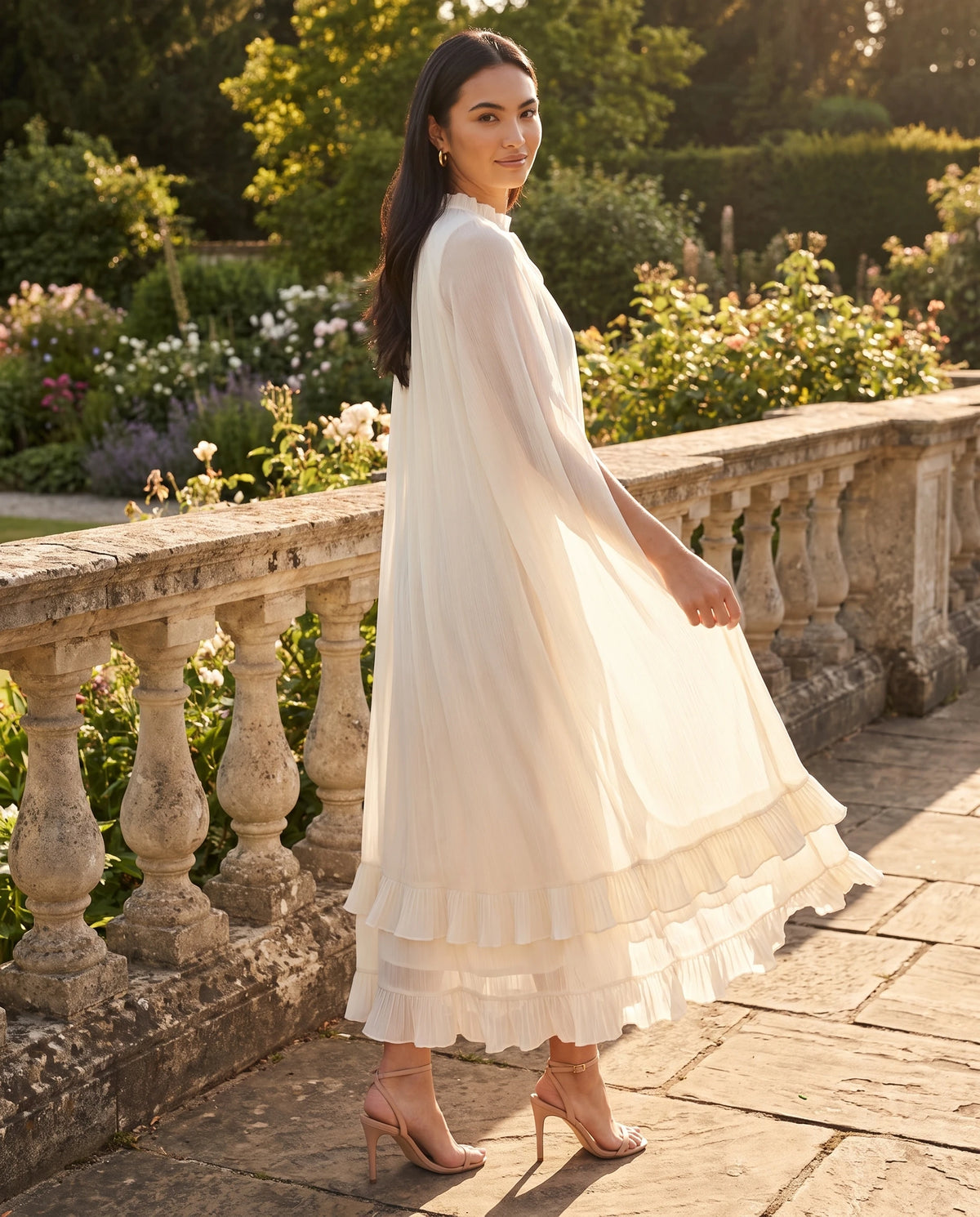 Robe longue cape en mousseline plissée Azalée en Ivoire, portée sur une terrasse en pierre au soleil