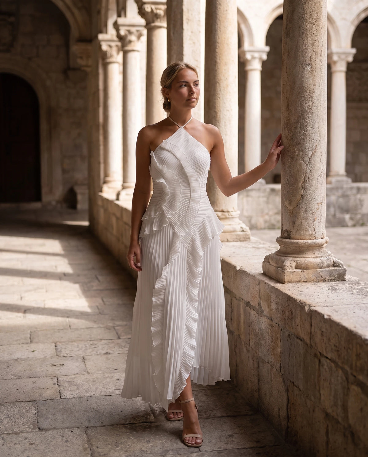 Robe longue halter plissée à volants Clélia en Blanc, portée dans une cour à colonnes de pierre