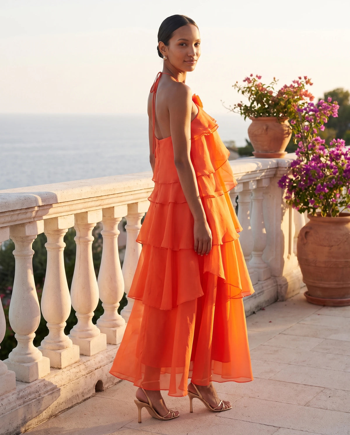 Langes Neckholder-Kleid aus Organza mit Stufenvolants Iliana in Orange, auf einer Terrasse bei Sonnenuntergang getragen