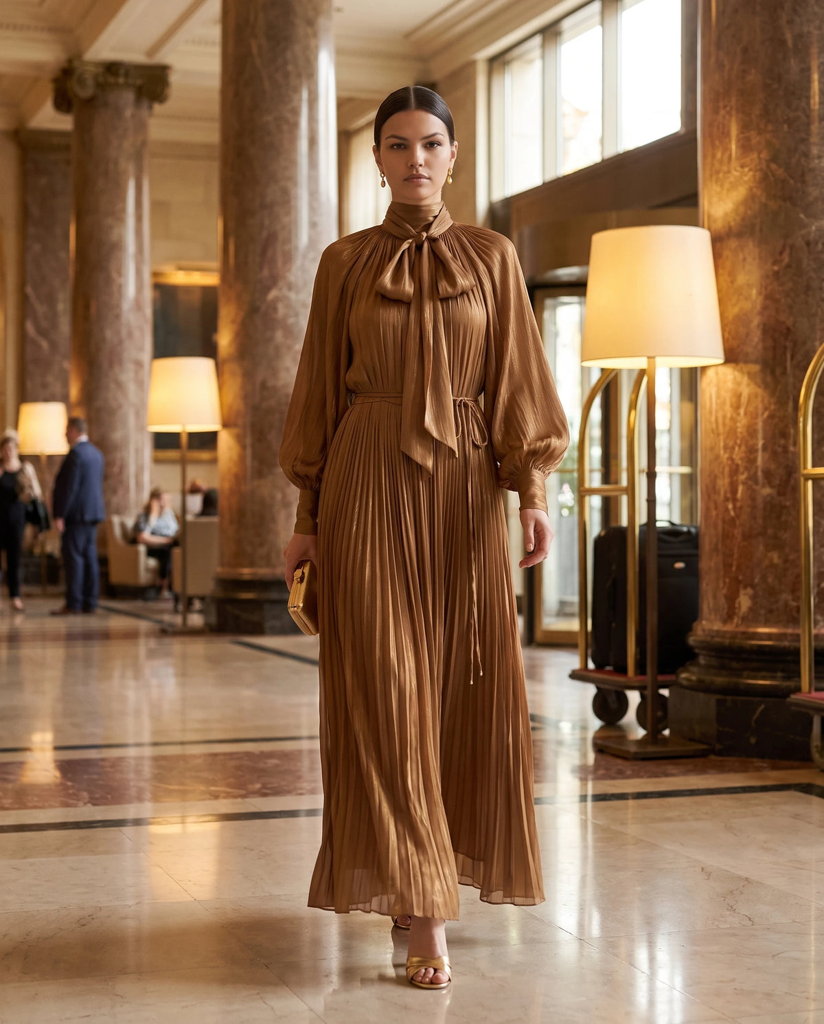 Robe longue plissée à col lavallière et manches chauve-souris Antonia en Bronze, portée dans le hall d'un grand hôtel
