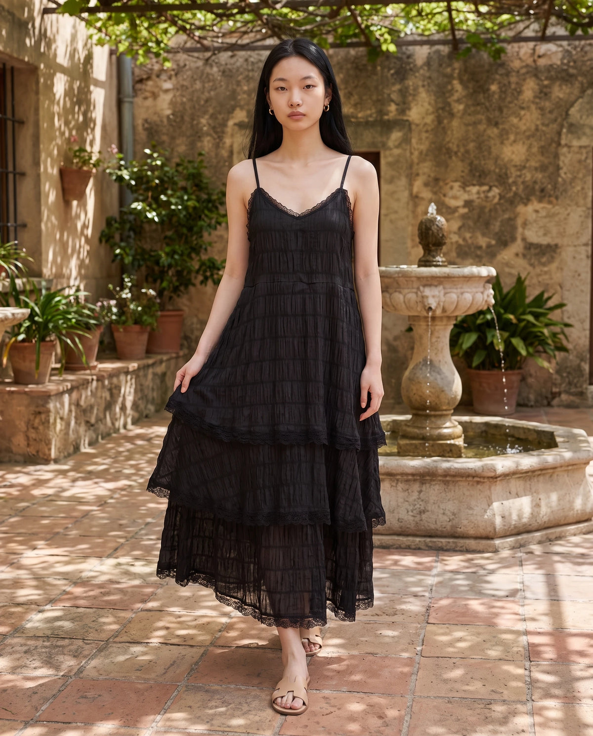 Robe longue à volants étagés et dentelle Elina en Noir, portée dans une cour ombragée à la fontaine de pierre