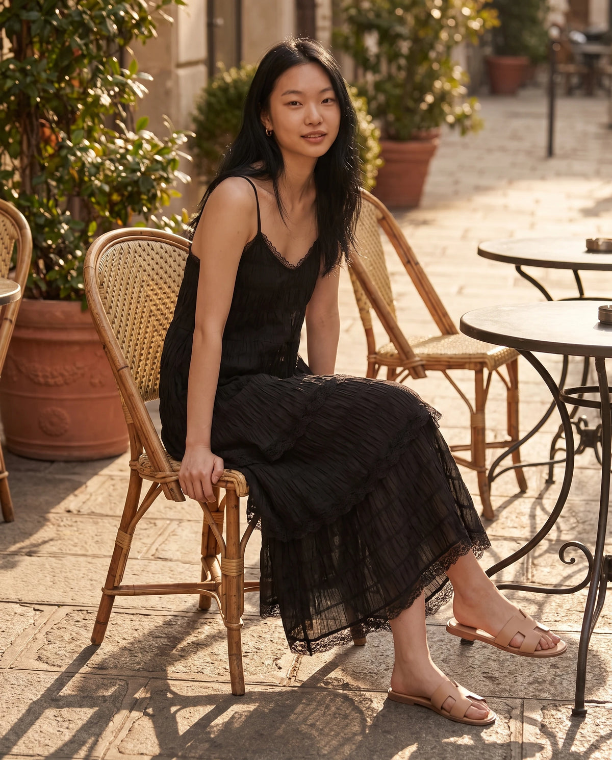 Robe longue à volants étagés et dentelle Elina en Noir, portée assise à la terrasse d'un café au soleil couchant
