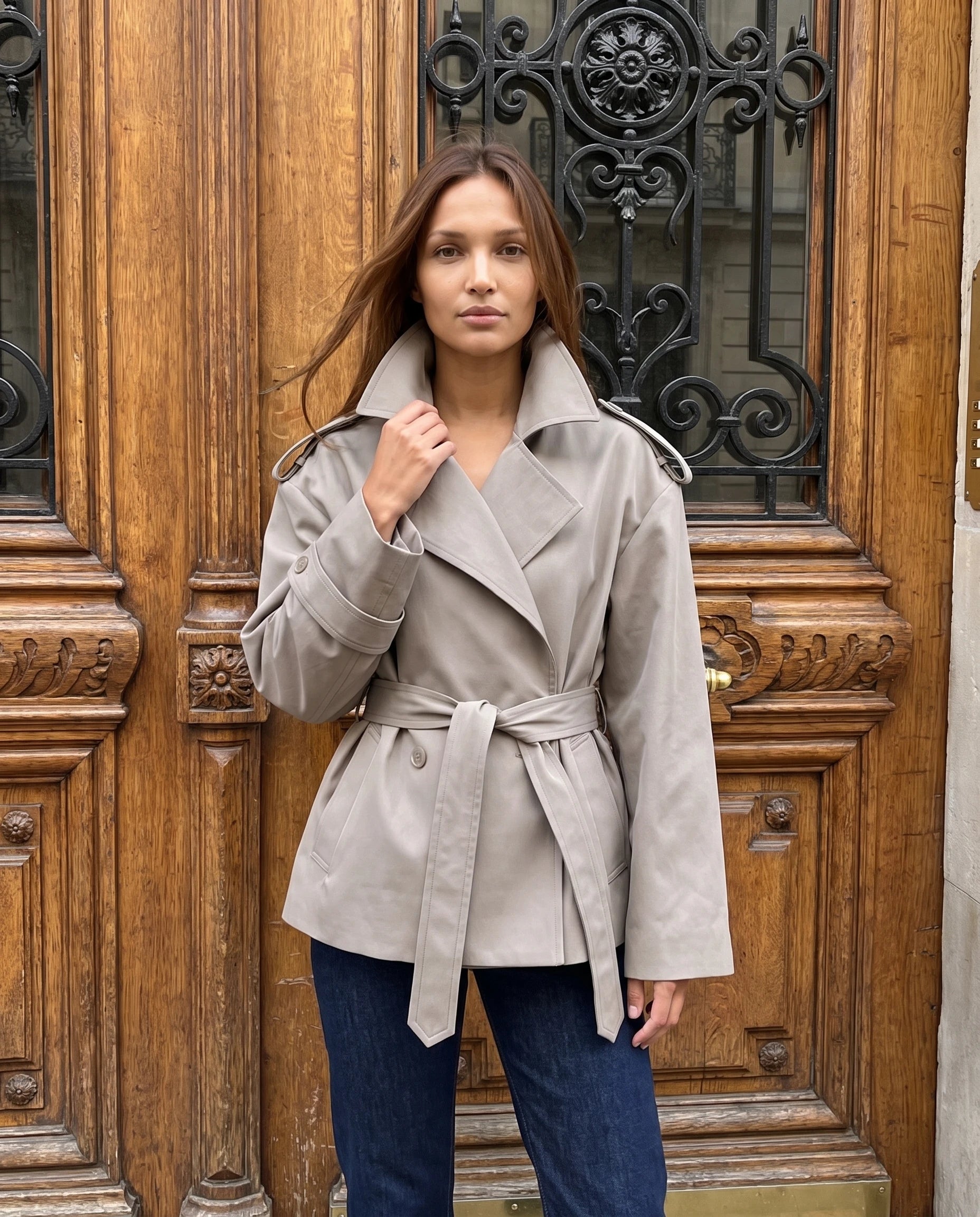 Trench court oversize taupe Bettina, devant une porte ancienne sculptée
