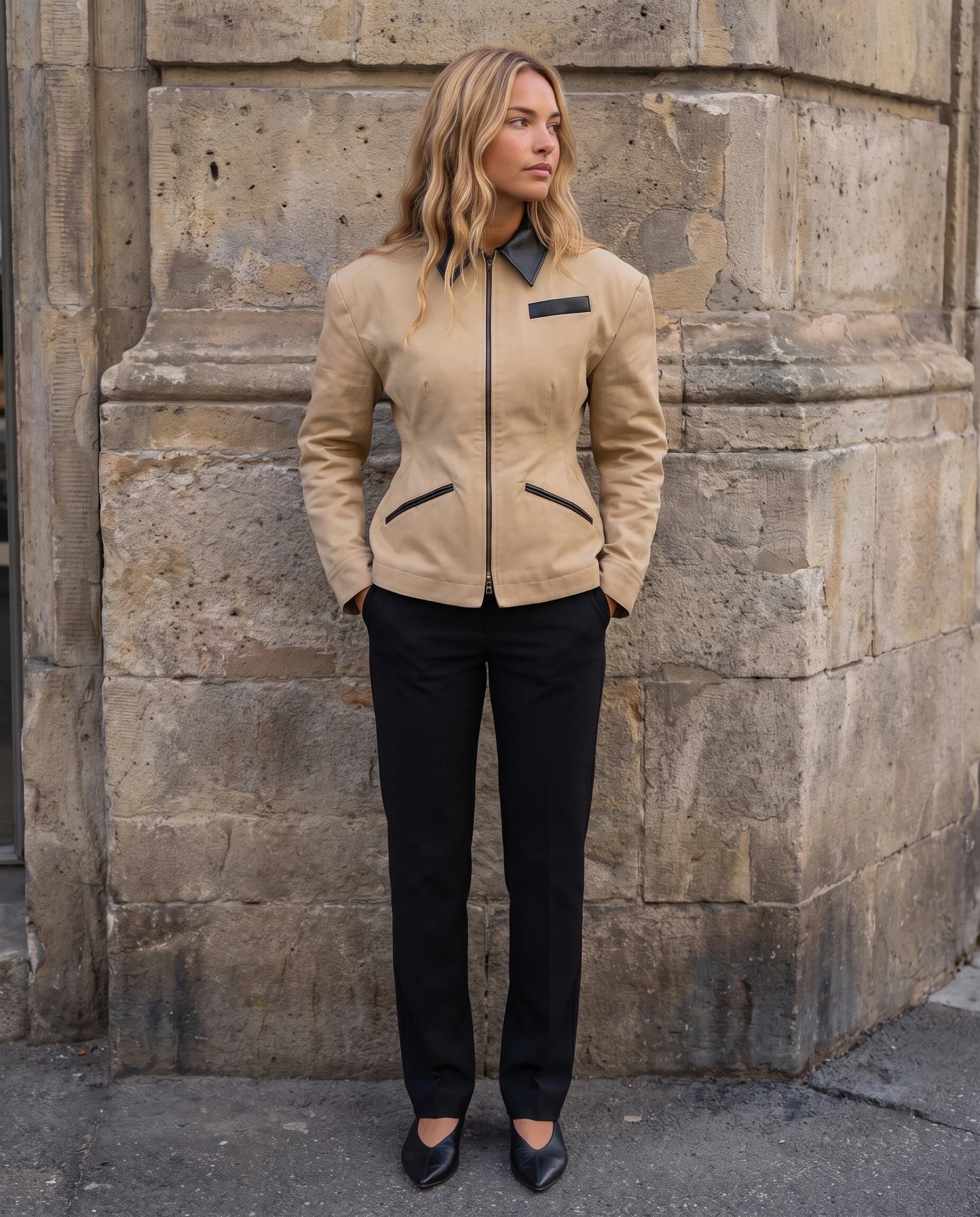 Veste cintrée à empiècements simili Charlotte, mains dans les poches devant un mur en pierre