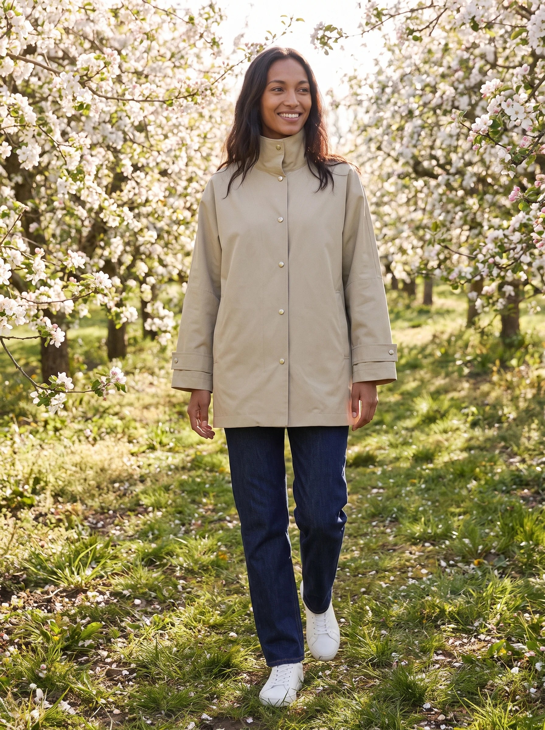 Veste mi-longue beige sable Mauricette, portée dans un verger de pommiers en fleurs