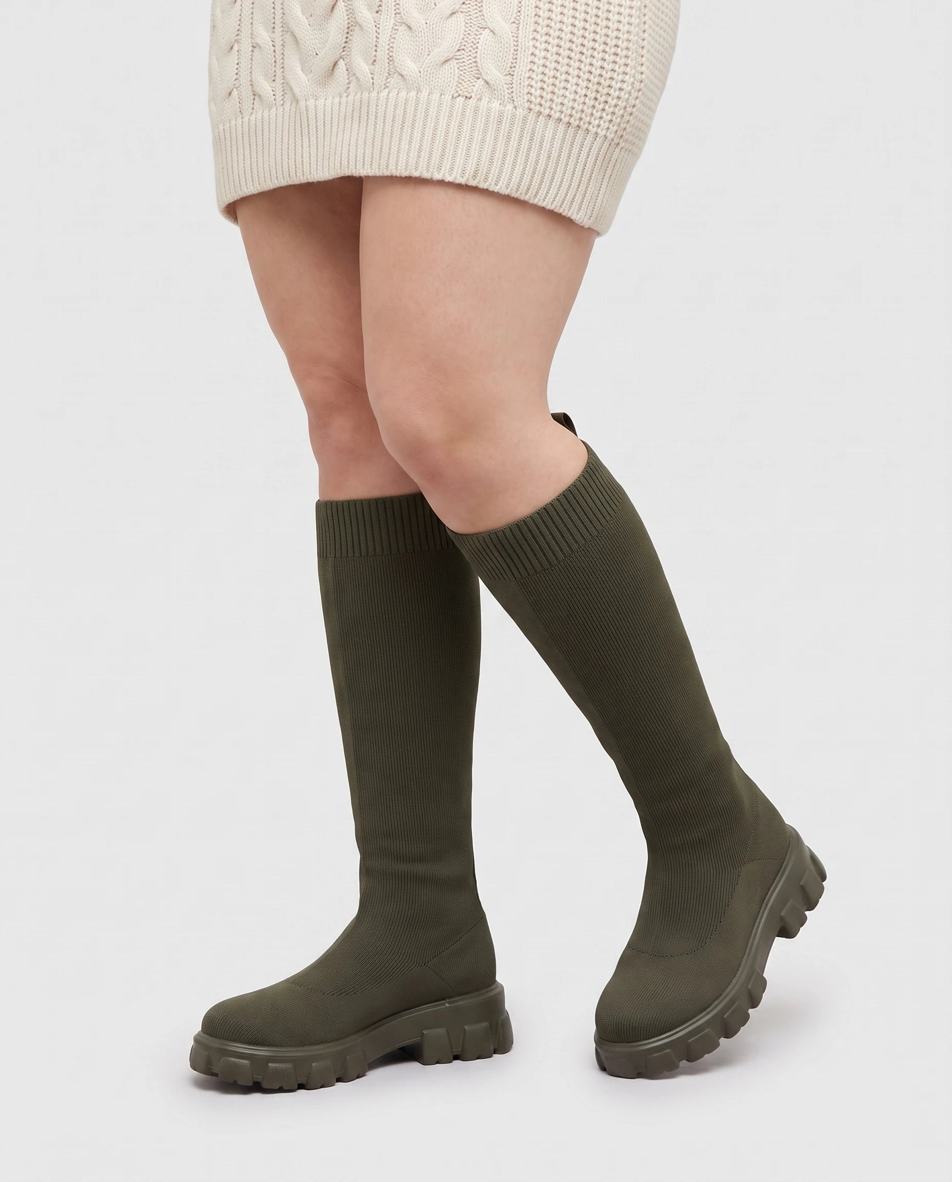 Bottes chaussettes hautes en maille Sylvette — Kaki, portées en plan rapproché avec une robe pull écrue