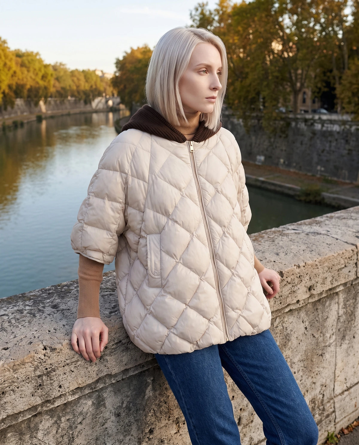 Cape matelassée à capuche tricot Stella en Écru, portée sur un pont de pierre en automne