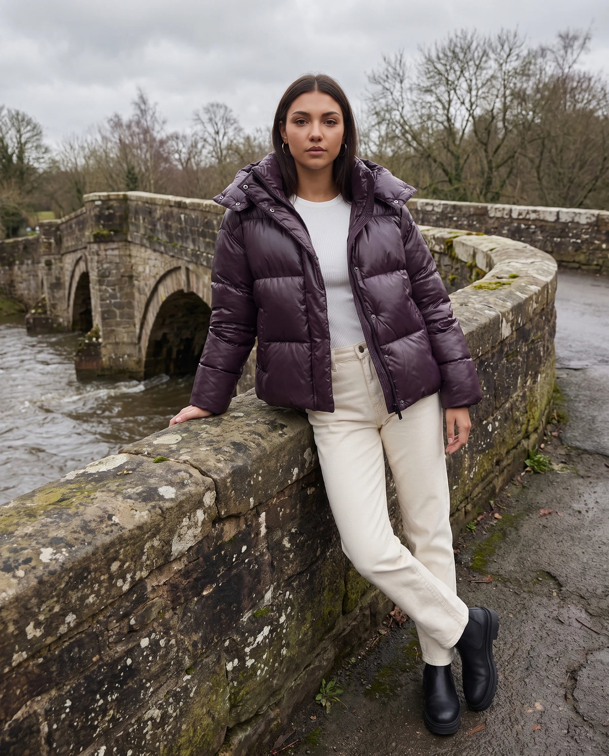 Doudoune brillante Tessa en Prune, portée sur un vieux pont de pierre