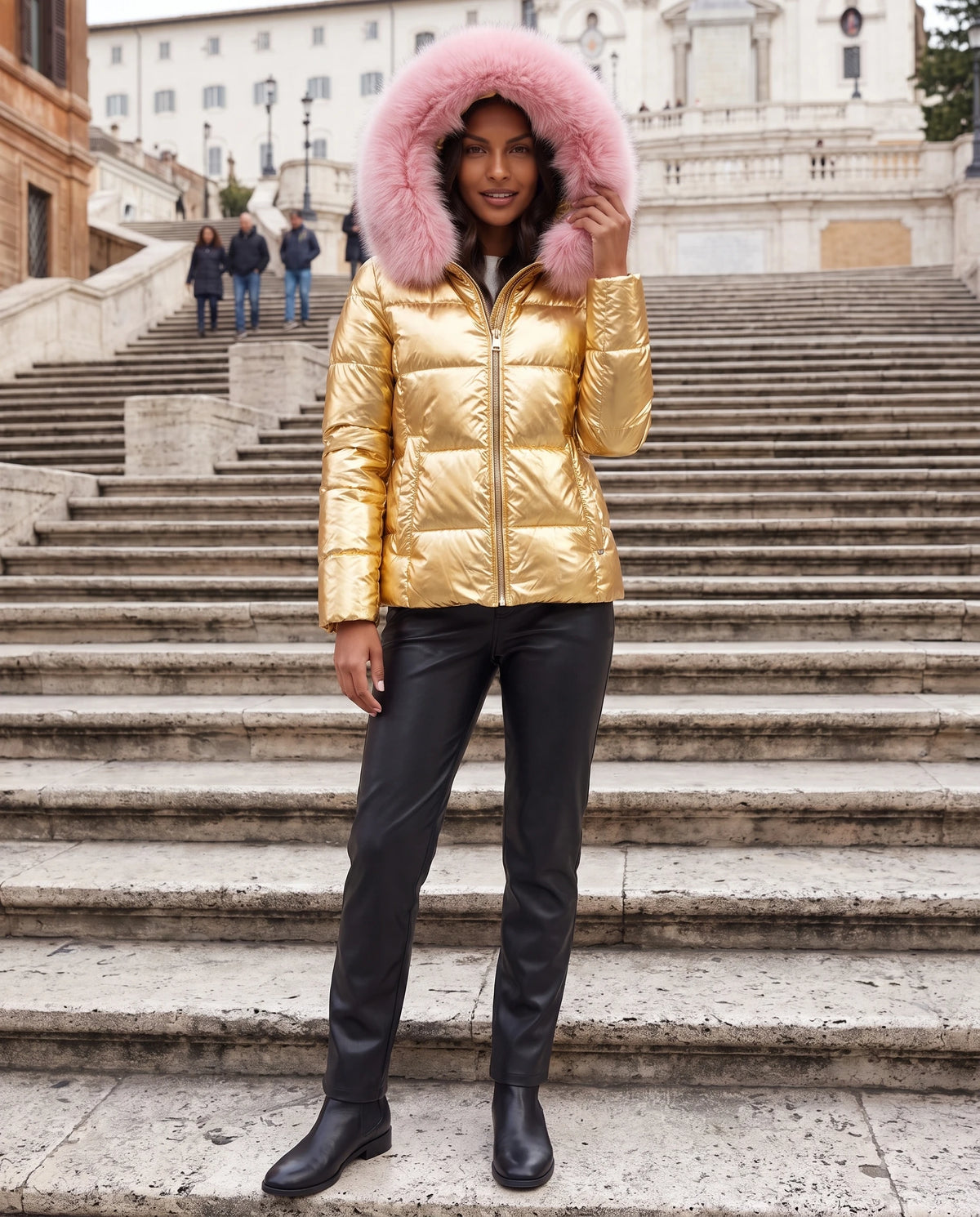 Kurze glänzende Steppjacke mit Kunstfellkapuze Victoria in Gold, getragen auf einer Steintreppe in der Stadt, Kapuze aufgesetzt