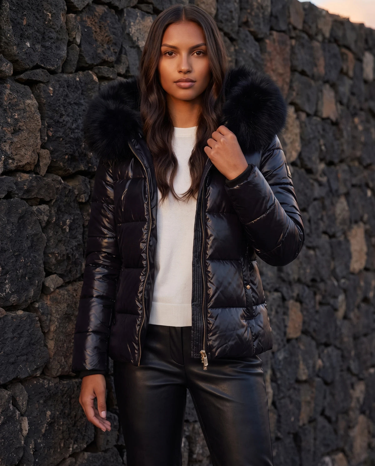 Kurze glänzende Steppjacke mit Kunstfellkapuze Victoria in Schwarz, getragen vor einer dunklen Steinmauer in der Dämmerung