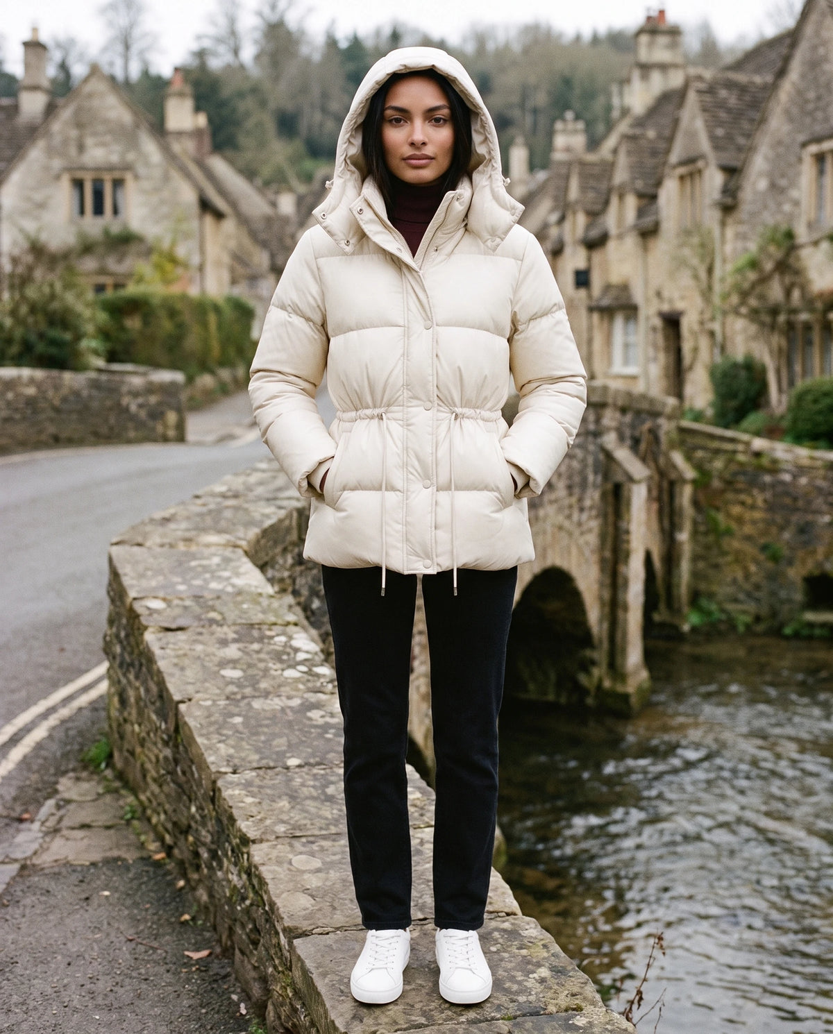 Kurze Steppjacke mit Kapuze und elastischer Taille Annick in Elfenbein, getragen mit Kapuze auf einer alten Steinbrücke im Dorf