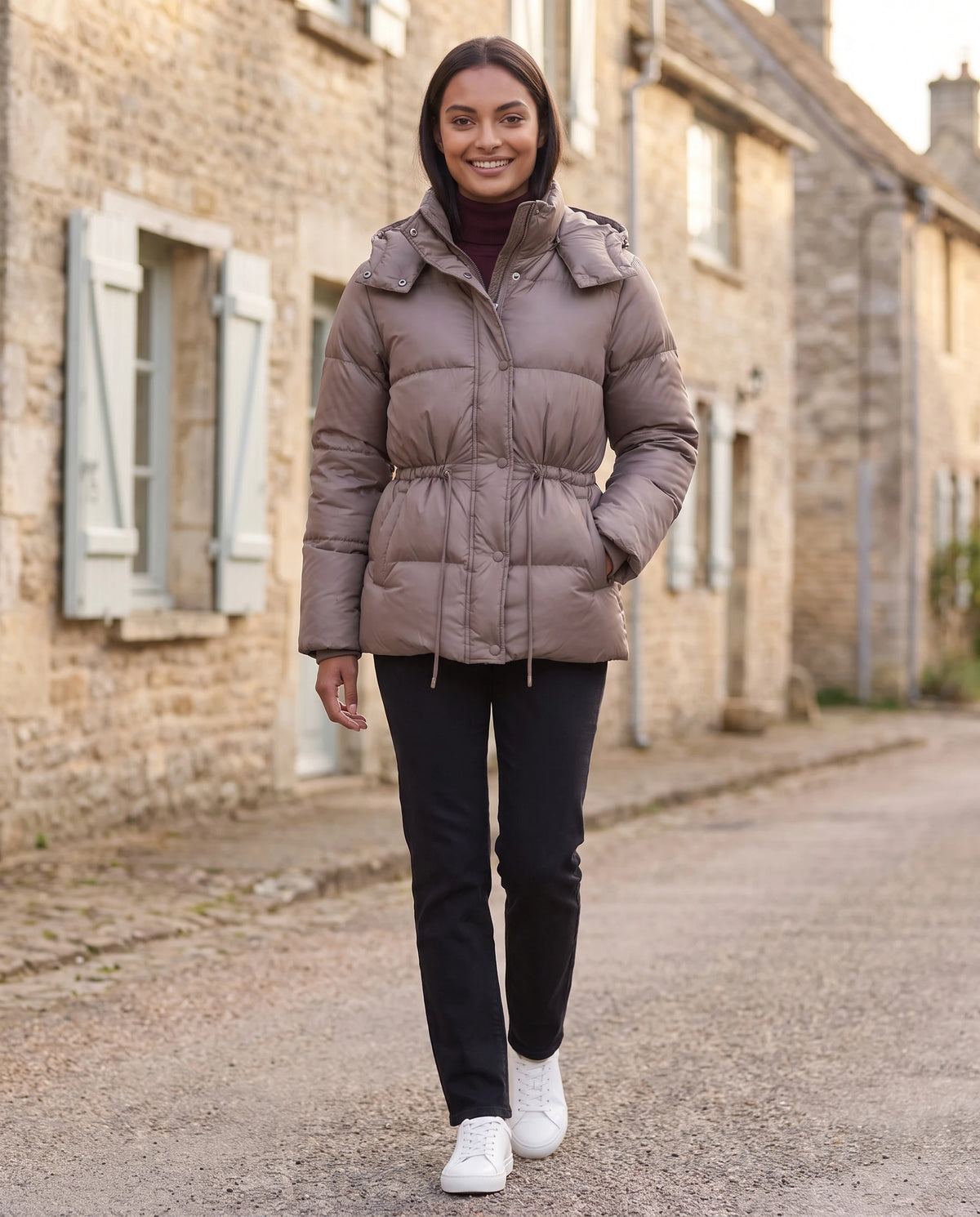 Kurze Steppjacke mit Kapuze und elastischer Taille Annick in Taupe, getragen in einer Dorfstrasse mit hellen Fensterläden