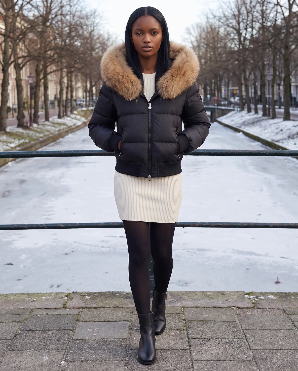 Kurze Steppjacke mit grosser Kunstfellkapuze Rebecca in Schwarz · naturfarbenes Fell, getragen auf einer Steinbrücke über einem zugefrorenen Kanal