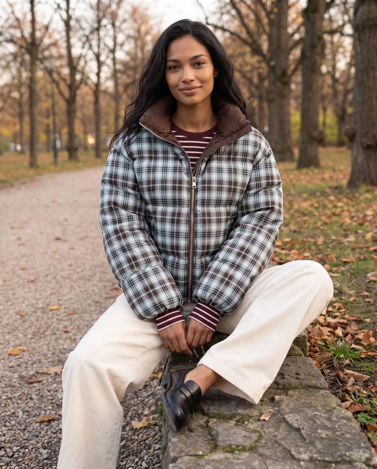 Doudoune courte réversible tartan à col montant Eve en Chocolat / Tartan, portée face tartan, assise sur un muret de pierre dans un parc en automne